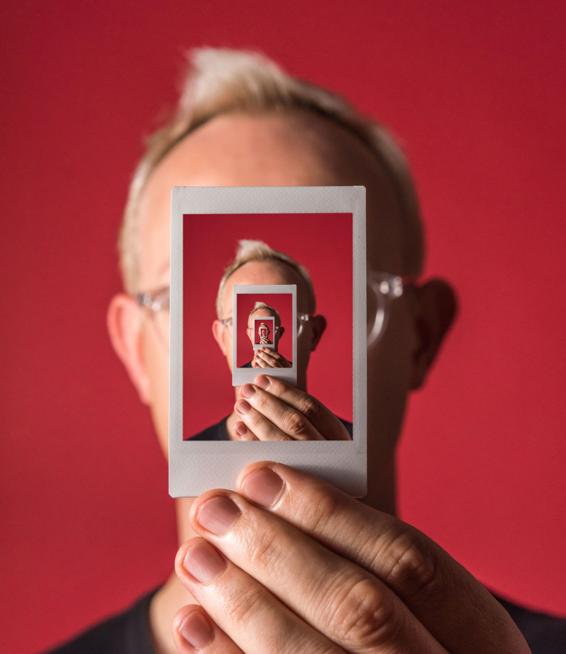 Red Polaroid A hand holding up a Polaroid-style picture in front of a face with the same image of a hand holding up a polaroid style picture infinitely repeating ever smaller and smaller. The background is red.